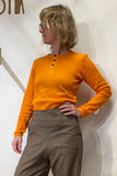A person with wavy blonde hair and glasses wears a BUTIK KUBIK Kubik cotton Henley Honey long-sleeve shirt and brown high-waisted pants, standing against a plain white wall with one hand on their hip.