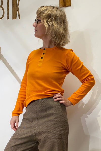A person with wavy blond hair and glasses stands sideways, wearing the BUTIK KUBIK Kubik cotton Henley Honey shirt and brown pants, one hand on their hip against a light-colored wall.