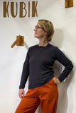 A person with blond hair and glasses stands confidently, hand on hip, wearing a BUTIK KUBIK Kubik cotton Henley in Graphite and orange pants. Wooden hooks and the word KUBIK are mounted on the white wall behind them.