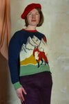 A person wearing the BUTIK KUBIK Palava Fred jumper navy alpine fox, a colorful organic cotton knit with a fox and mountain design, stands against a textured wall looking slightly upwards.