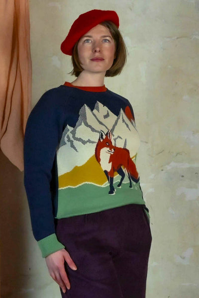 A person wearing the BUTIK KUBIK Palava Fred jumper navy alpine fox, a colorful organic cotton knit with a fox and mountain design, stands against a textured wall looking slightly upwards.