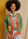 A curly-haired woman smiles in a BUTIK KUBIK Palava George cardigan ice cream, featuring green and coral hues with colorful ice cream cone patterns, layered over a plaid shirt and striped skirt against beige and yellow fabric.