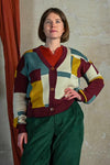 A woman with short brown hair stands against a textured wall, smiling slightly with one hand on her hip. She wears a BUTIK KUBIK Palava George plum boxes cardigan over a red top and green corduroy pants.