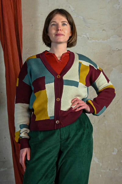 A woman with short brown hair stands against a textured wall, smiling slightly with one hand on her hip. She wears a BUTIK KUBIK Palava George plum boxes cardigan over a red top and green corduroy pants.