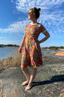 A woman stands barefoot on a rocky shore in the Kubik dress Twist Golden flowers by BUTIK KUBIK, made from OEKOTEX-certified fabric. The 50s-inspired floral dress shines under blue skies beside calm water and reeds.