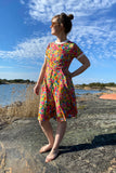 A woman stands barefoot on a rocky shore in the Kubik dress Twist Golden flowers by BUTIK KUBIK, made from OEKOTEX-certified fabric. The 50s-inspired floral dress shines under blue skies beside calm water and reeds.