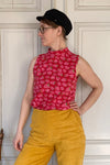 A person in a black hat and the Kubik top cineast Tarja from BUTIK KUBIK, featuring a 60s inspired floral pattern, stands against a white paneled wall, wearing yellow pants and gazing to the side.