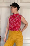 A person in a black hat and the Kubik top cineast Tarja from BUTIK KUBIK, featuring a 60s inspired floral pattern, stands against a white paneled wall, wearing yellow pants and gazing to the side.