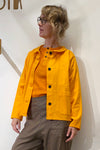 A person with wavy blond hair and glasses wears the BUTIK KUBIK Kubik Overshirt vintage yellow over an orange top and brown pants, standing against a white wall and looking to the side.