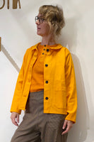 A person with wavy blond hair and glasses wears the BUTIK KUBIK Kubik Overshirt vintage yellow over an orange top and brown pants, standing against a white wall and looking to the side.