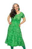 A woman with long gray hair smiles and poses in the Cissi och Selma Hedvig dress Ängsblom Grön by BUTIK KUBIK, paired with green earrings and tights, against a white background.