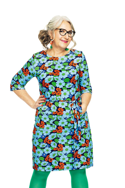 A confident woman with gray hair and glasses stands with one hand on her hip, wearing the Cissi och Selma dress Doris Humlesurr blå by BUTIK KUBIK in an A-line ekologisk bomullstrikå fabric with a 60-talet floral pattern, complemented by green tights against a white backdrop.