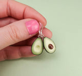 A hand with pink nail polish holds a pair of Becca design Avokado earrings by BUTIK KUBIK—one with a brown seed and one without—against a light green background.