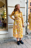 A woman stands on a cobblestone sidewalk, smiling and looking to the side. She wears the Kubik dress Cirrus by BUTIK KUBIK with black shoes. A shop window and hanging dress are visible in the background.
