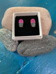 Törmi earrings Verho mini burgundy by BUTIK KUBIK—oval Finnish birch plywood earrings with a pink top and black-and-white checkered bottom—are displayed in a white box on black velvet among smooth stones atop blue wood.
