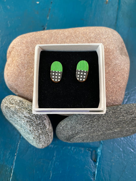 Törmi earrings Verho mini green by BUTIK KUBIK: oval Finnish birch earrings with green tops, black-and-white checkered bottoms, eco-packaged and displayed on smooth stones against a blue wooden background.