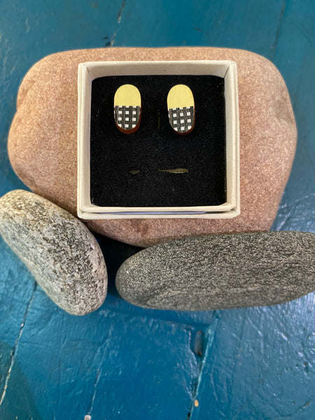 The Törmi earrings Verho mini yellow by BUTIK KUBIK are oval-shaped Finnish Birch plywood earrings with yellow tops and black-and-white checkered bottoms, shown in a small box on three smooth stones against a blue wooden background.