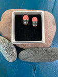 A pair of Törmi earrings Verho mini coral by BUTIK KUBIK, featuring pink tops and black-and-white checkered Finnish birch plywood bottoms, rest in a small box on a stone amid pebbles on a blue wooden surface.