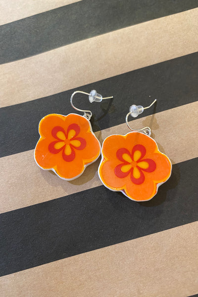 Saved & remade earrings by BUTIK KUBIK: silverplated, orange flower-shaped, with yellow and red details, crafted from upcycled porcelain. Shown on a striped black and tan surface.