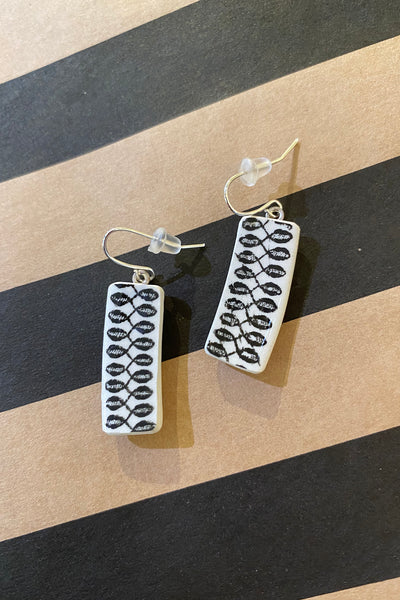 A pair of rectangular white Saved & Remade earrings by BUTIK KUBIK, made from recycled porcelain with black leaf patterns, rests on diagonal tan and black stripes—elegant upcycled style for your individuality.