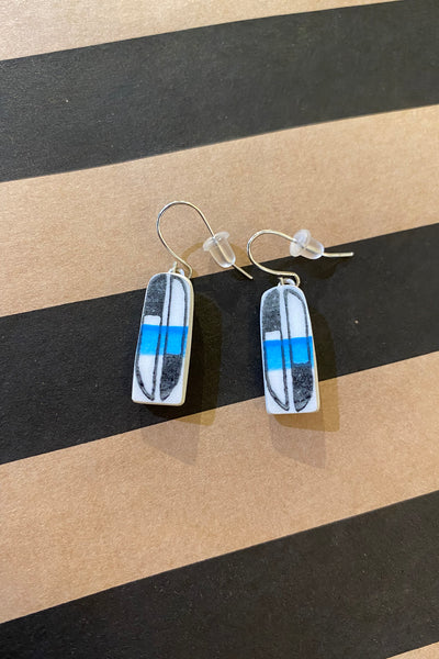 A pair of Saved & remade earrings by BUTIK KUBIK, made from återbrukat porslin, with black outlines and blue horizontal stripes in the center, lies on a brown surface with wide black diagonal stripes.