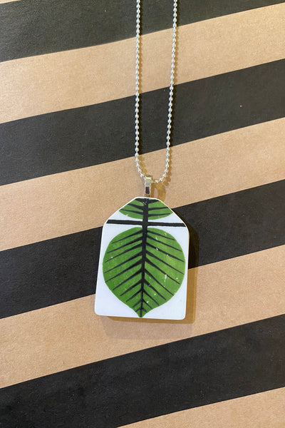 The Saved & remade necklace Porslin by BUTIK KUBIK features a green leaf with black veins on återbrukat porslin, set against white. Elegant silverpläterad kedja. Displayed on a tan and black striped surface.