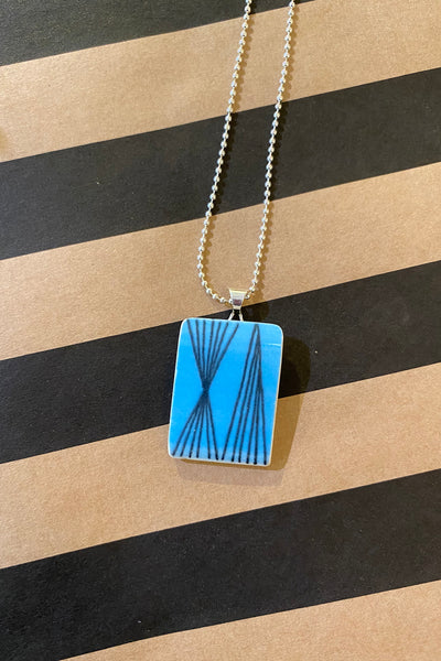 The Saved & remade necklace Porslin by BUTIK KUBIK features a silverpläterad kedja with a square pendant of återbrukat porslin, showcasing a bright blue background and black geometric lines, displayed on a striped brown and black surface.