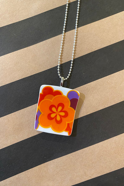 The Saved & remade necklace Porslin by BUTIK KUBIK features a square pendant with an orange, yellow, and red flower design made from recycled porcelain on a silver-plated chain, shown against a tan and black striped background.