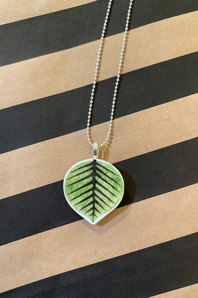 Saved & remade necklace Porslin från BUTIK KUBIK: Ett silverpläterat halsband med bladformat, grönt och svartmönstrat porslinshänge, framhävt på en brun bakgrund med diagonala svarta ränder.