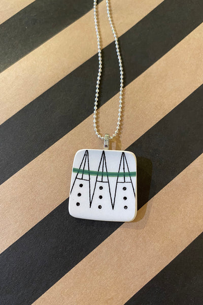 The Saved & remade necklace Porslin by BUTIK KUBIK features a square pendant made from återbrukat porslin with black triangles, dots, and a green line on silverpläterad kedja, shown on a diagonally striped background.
