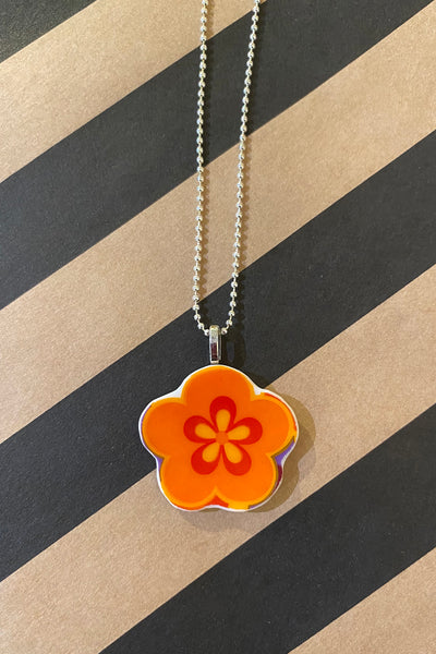 The Saved & Remade necklace Porslin by BUTIK KUBIK is a silverplated chain with a flower-shaped orange pendant featuring a red floral design, shown on a diagonal black and tan striped background.