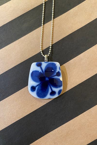 The Saved & remade necklace Porslin by BUTIK KUBIK features blue florals on återbrukat porslin, hanging from a silverpläterad kedja, and is displayed against wide brown and black diagonal stripes.
