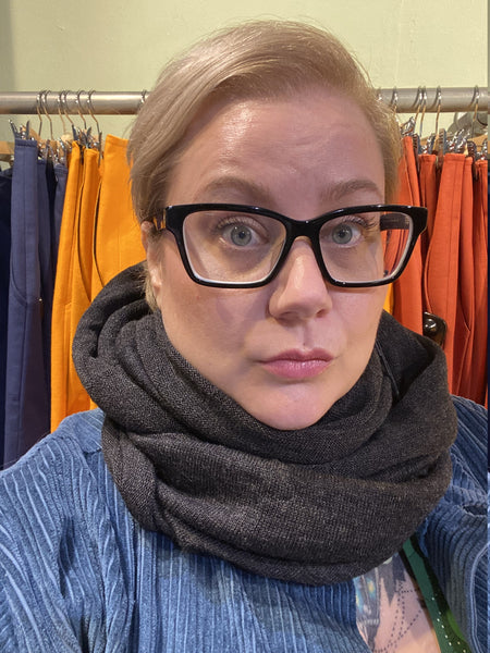 A person with short blonde hair, black glasses, a blue top, and the Mariedal Tubsjal Grafit from BUTIK KUBIK stands before a clothing rack displaying orange and blue pants, wearing a neutral expression.
