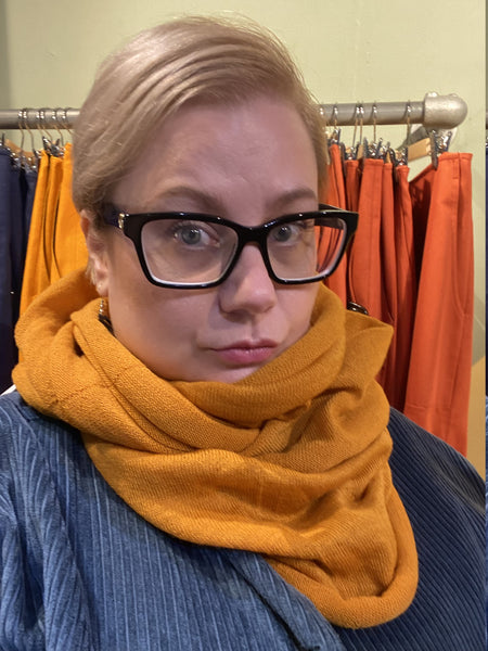A blond person in black glasses wears the BUTIK KUBIK Mariedal Tubsjal Saffran and a blue textured top, standing before orange and navy garments hanging on a rack.
