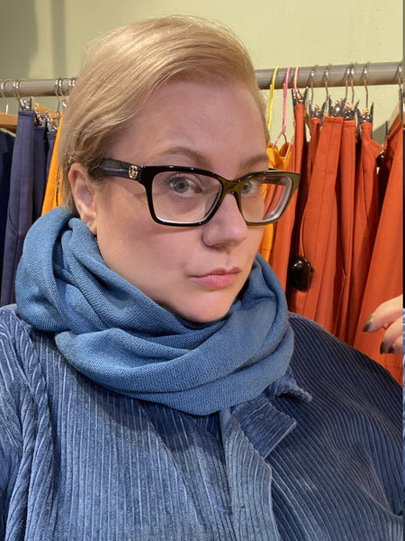 A person with short blond hair and black glasses wears the Mariedal Tubsjal Petrol scarf by BUTIK KUBIK and a blue textured jacket, standing before a rack filled with colorful orange, red, and yellow clothes.