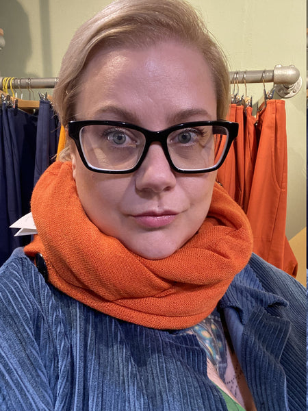 A person with short blonde hair and black glasses wears the Mariedal Tubsjal Orange from BUTIK KUBIK and a blue jacket, standing indoors by a rack displaying orange and blue garments.