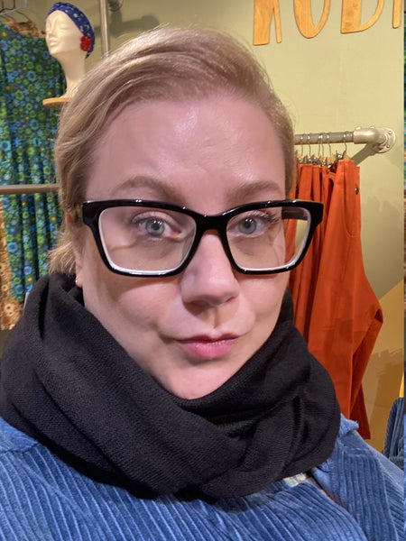 A person with light hair and glasses, wearing the Mariedal Tubsjal Svart from BUTIK KUBIK and a blue top, stands indoors before orange clothing and a mannequin with a floral headband.