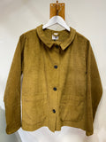 The BUTIK KUBIK Kubik corduroy overshirt in olive, featuring black buttons and two large front pockets, hangs on a white hanger against a white wall.