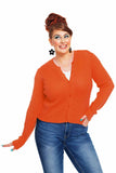 A smiling woman with bright makeup and a red updo wears the vibrant orange Cissi och Selma cardigan Apelsin from BUTIK KUBIK, paired with blue jeans, a white top, and large black flower earrings, posing confidently against a white background.
