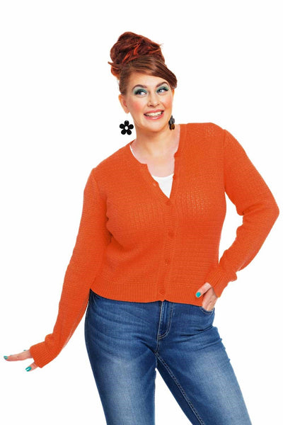 A smiling woman with bright makeup and a red updo wears the vibrant orange Cissi och Selma cardigan Apelsin from BUTIK KUBIK, paired with blue jeans, a white top, and large black flower earrings, posing confidently against a white background.