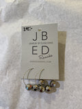 A pair of JBED Jewelry By Elina Dahl - Remake Örhängen from BUTIK KUBIK features faceted metallic beads made from återvunna pärlor, displayed on a labeled card with "295:-" handwritten, set atop crumpled white tissue paper.