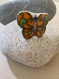 The Saved & remade ring porslin Fjäril from BUTIK KUBIK is a colorful, butterfly-shaped silverpläterad ring crafted from återbrukat porslin, featuring justerbar storlek and displayed on a speckled stone with a gray stone backdrop.