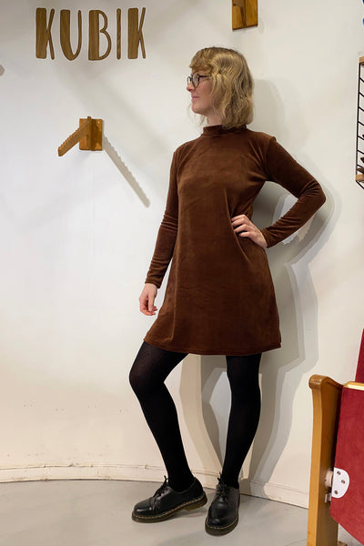 A person in glasses and the BUTIK KUBIK Kubik dress cinema velour with black tights and shoes stands in a minimalist room featuring “KUBIK” on the wall behind them, looking to the side with one hand on their hip.