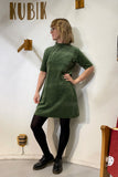A person wearing glasses and the BUTIK KUBIK "Kubik dress cinema velour," a 60s-inspired green A-line long sleeve dress, with black tights and shoes, stands hand-on-hip before a white wall with KUBIK and wooden decorations.