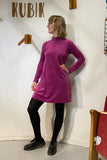 A person with short blond hair and glasses wears the BUTIK KUBIK "Kubik dress cinema velour," a 60's inspired magenta A-line long sleeve dress, paired with black tights and shoes, posing under wooden KUBIK letters against a white wall.