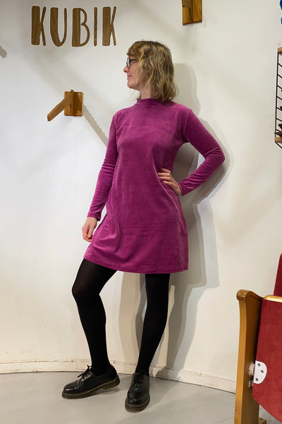 A person with short blond hair and glasses wears the BUTIK KUBIK "Kubik dress cinema velour," a 60's inspired magenta A-line long sleeve dress, paired with black tights and shoes, posing under wooden KUBIK letters against a white wall.