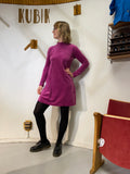 A person wearing the Kubik dress cinema velour by BUTIK KUBIK in magenta, with black tights and shoes, stands indoors near a red chair next to a wall displaying "KUBIK" and wooden hooks. Made in Sweden.