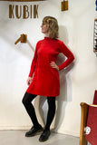 A woman with wavy blonde hair, glasses, and black tights stands against a white wall, wearing the BUTIK KUBIK Kubik dress cinema velour with black shoes. The word KUBIK appears above her on the wall.