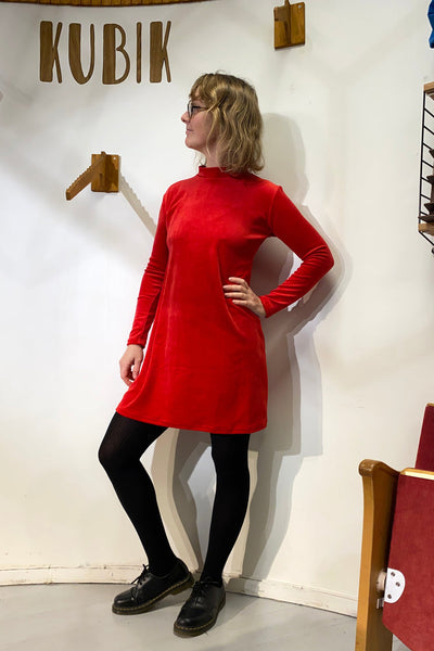 A woman with wavy blonde hair, glasses, and black tights stands against a white wall, wearing the BUTIK KUBIK Kubik dress cinema velour with black shoes. The word KUBIK appears above her on the wall.