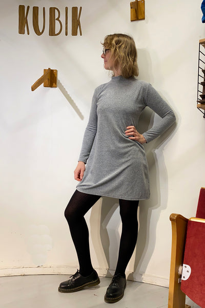 A person with shoulder-length blonde hair and glasses stands against a white wall beneath the KUBIK sign, wearing the BUTIK KUBIK Kubik dress cinema velour in grey with black tights and shoes, hand on hip and looking to the side.